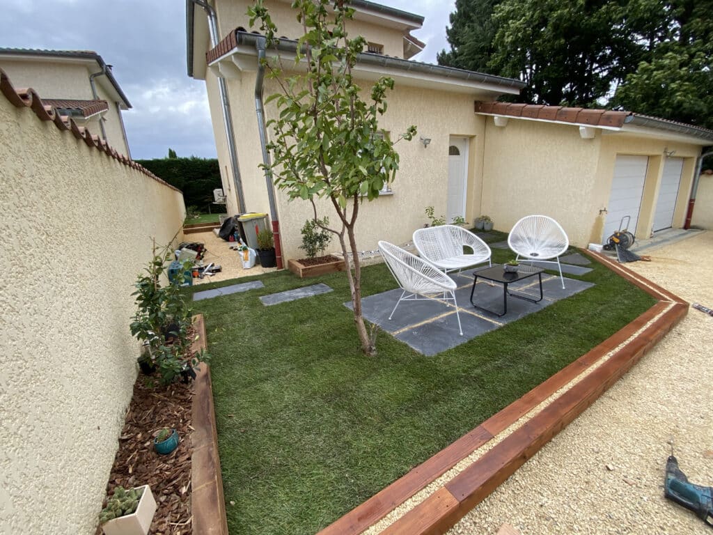 Petit jardin moderne avec chaises blanches, terrasse en ardoise, pelouse verte et bordure en bois devant une maison.