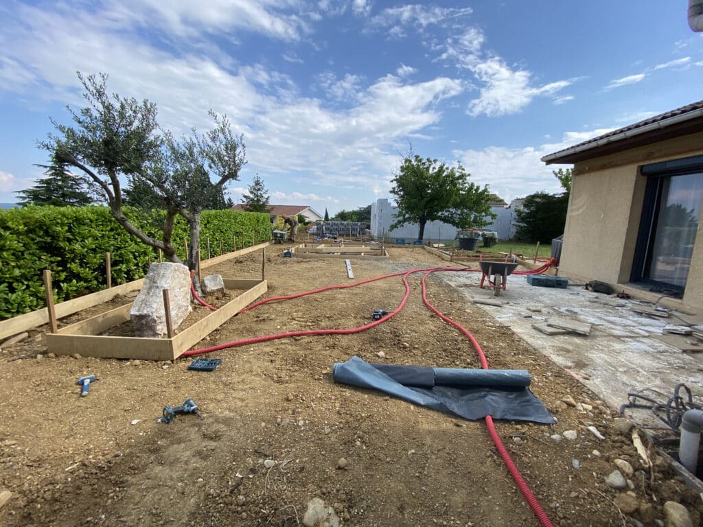 Chantier d'aménagement paysager avec coffrage de terrasse, olivier et gaines rouges dans un jardin ensoleillé.