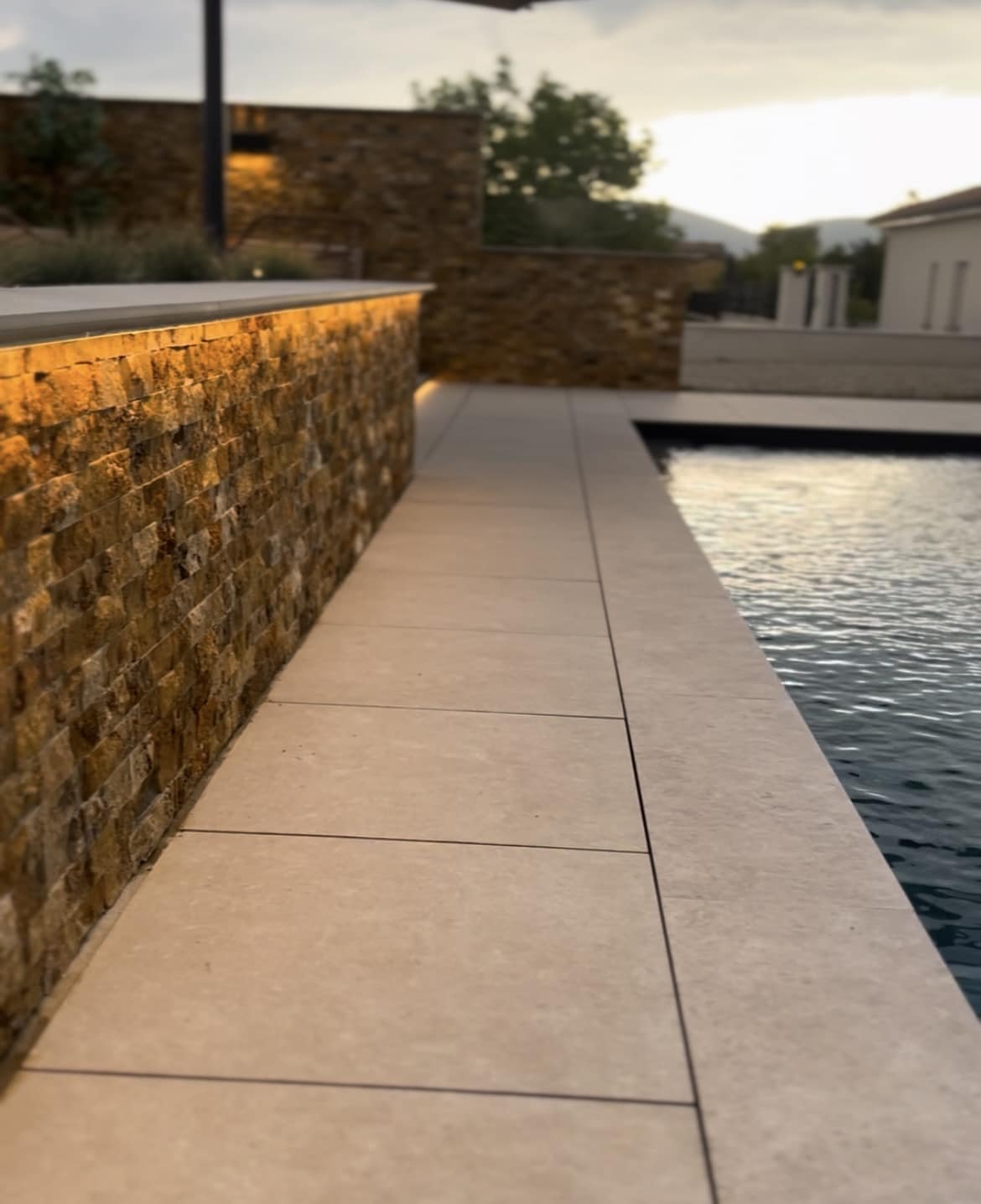 Aménagement piscine moderne avec mur en pierre et LED Margelle de piscine éclairée par un bandeau LED sous un mur en pierre naturelle en soirée.