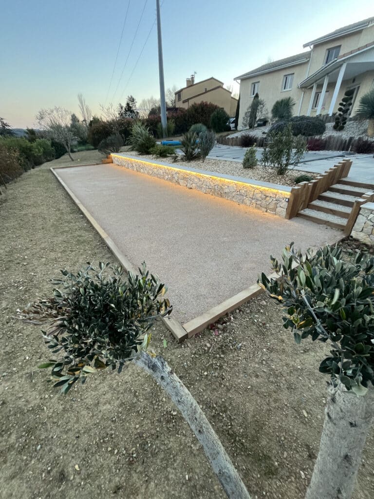 Terrain de pétanque avec muret en pierre éclairé, escalier bois et oliviers dans un jardin paysager au crépuscule.