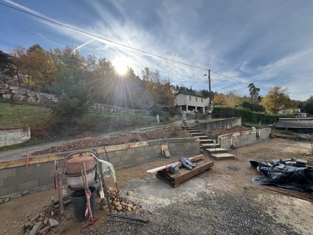 Chantier de construction : mur en parpaings, bétonnière et escaliers extérieurs sous un soleil d'automne éclatant.