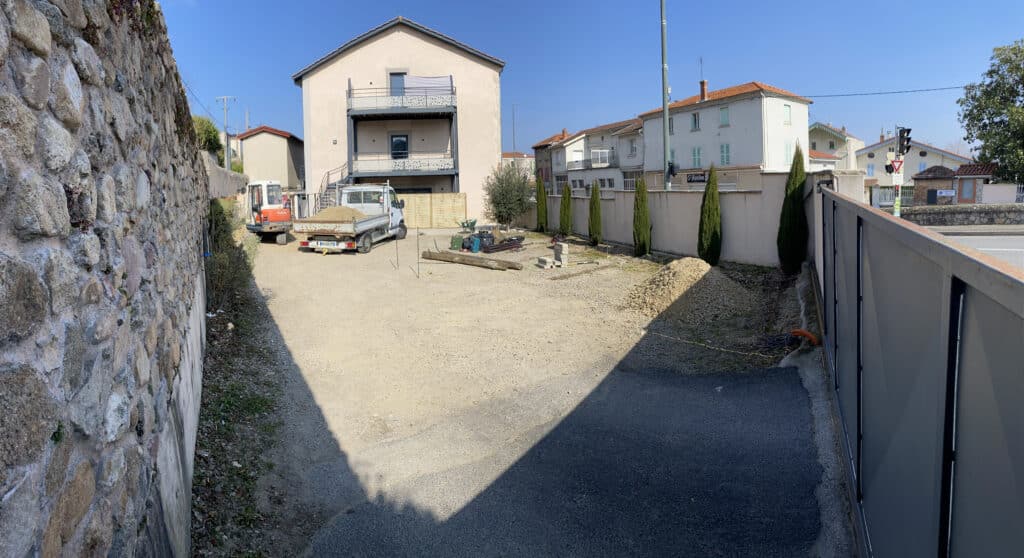 Chantier de terrassement avec camion, mini-pelle et tas de gravier devant une maison moderne sous un ciel bleu dégagé.