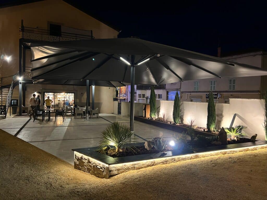 Terrasse nocturne moderne avec grand parasol gris éclairé, jardinières en pierre illuminées et mobilier de jardin.