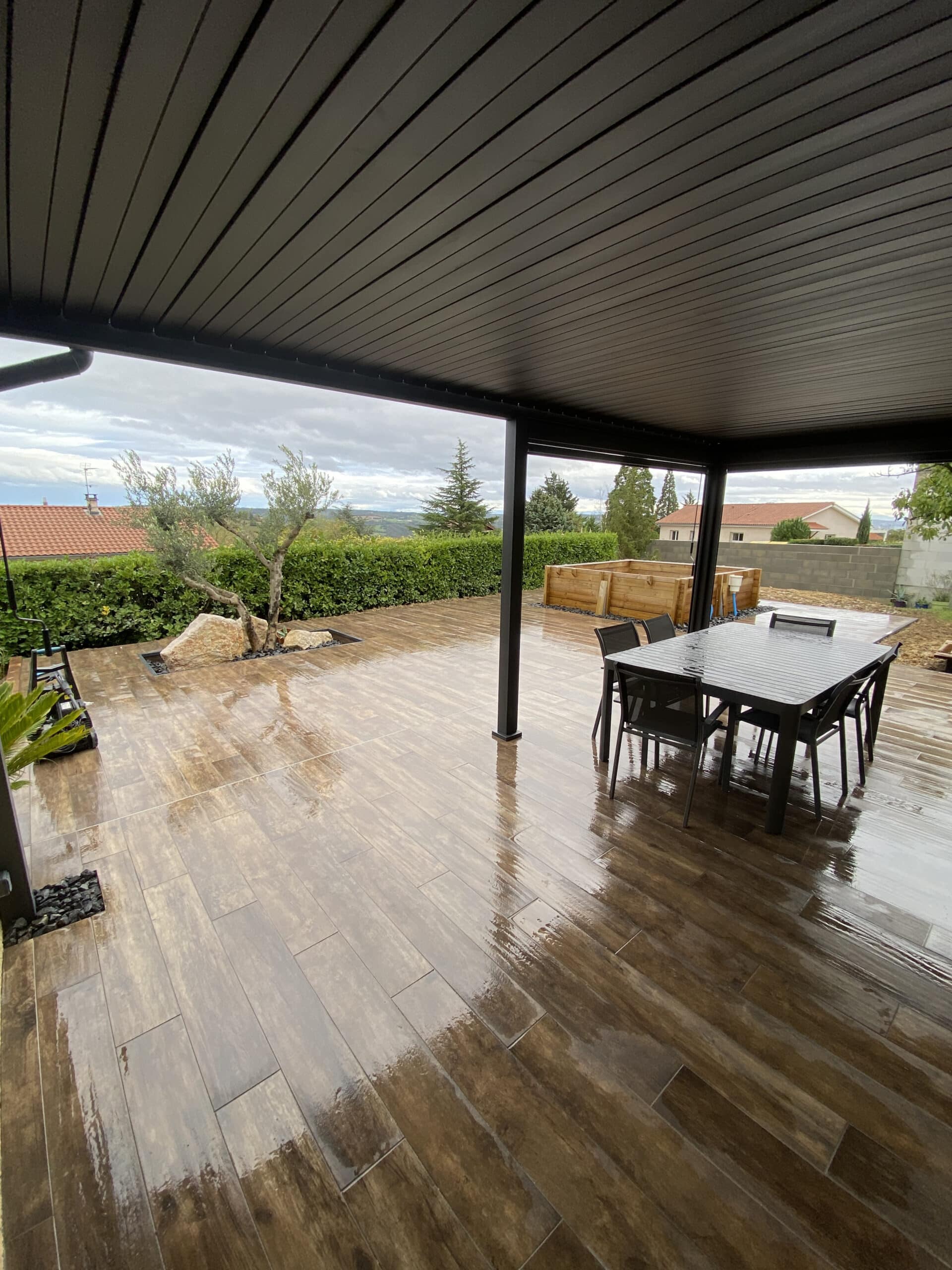 Pergola bioclimatique et terrasse en carrelage aspect bois Terrasse moderne avec pergola noire, carrelage aspect bois et mobilier de jardin par temps de pluie.