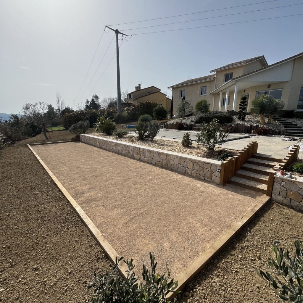 Terrain de pétanque avec muret en pierre et bois Terrain de pétanque en sable avec muret en pierre et escalier en bois dans un jardin paysager ensoleillé.