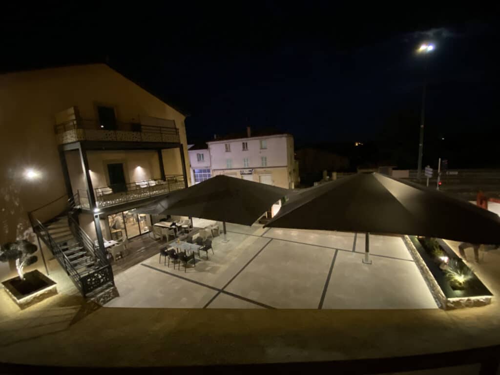 Terrasse de restaurant nocturne, grands parasols sombres, mobilier d'extérieur et éclairage tamisé sur cour pavée.